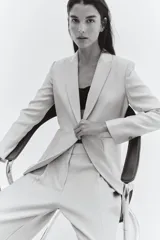 Blazer color beige de corte entallado, con solapas de pico, cierre de un botón, hombreras, bolsillos delanteros de parche, puños con abertura y botón, y abertura trasera.