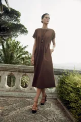 Vestido midi color marrón confeccionado en hilatura con mezcla de algodón y lino. Tiene cuello redondo, manga corta caída y detalle de pliegues en la cintura. Cierre en la espalda con abertura y botón.