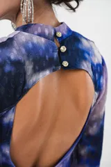 Vestido largo con estampado abstracto en tonos azules, violetas y grises. Tiene cuello mao con abertura en forma de gota, mangas largas acampanadas con detalle de botón en los puños, espalda abierta, tajo frontal y flecos en el ruedo.