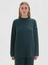 Buzo tejido color verde oscuro, con cuello subido tipo polera, diseño oversized y largo con pequeños cortes laterales. Presenta un patrón de trenzas y líneas verticales.