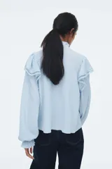 Blusa celeste de manga larga con textura arrugada, cuello alto con volados y abertura frontal con lazos para atar. Presenta canesú con volados y detalles de encaje calado en el pecho y hombros. Los puños terminan con elástico y volado.