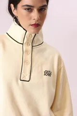 Buzo color crema con cuello alto con botones y ribete negro en cuello, puños y ruedo.