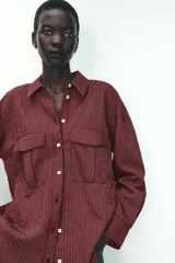 Camisa color borgoña a rayas verticales finas, de corte recto, con cuello solapa y manga larga. Tiene dos bolsillos de parche con solapa en el pecho y cierre frontal con botones.