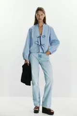 Camisa celeste con rayas verticales marrones y blancas, de cuello redondo con escote pico y manga larga abullonada. Cierre frontal con lazos con pespuntes marcados.