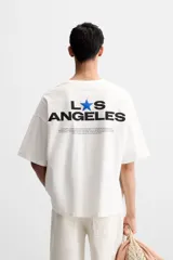 Camiseta blanca de corte holgado, cuello redondo y manga corta. Presenta un pequeño estampado de una estrella azul y un texto en negro en el frente.