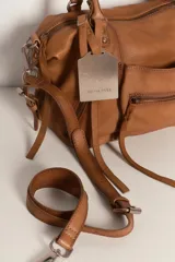 Bolso de mano tipo satchel o tote pequeño de cuero marrón con efecto lavado y apariencia vintage. Presenta dos asas superiores, múltiples cierres externos con tiradores de cuero y una etiqueta colgante metálica con logo.