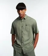 Camisa verde oliva de corte relajado, confeccionada en cáñamo y algodón. Tiene cuello inglés, mangas cortas, abotonadura frontal y bolsillo en el pecho con etiqueta de la marca.