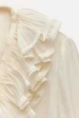 Blusa color crudo confeccionada en hilatura con mezcla de lyocell. Escote pico ajustable con cordones, manga larga con volumen acabada en elástico y detalle de volantes en el mismo tejido.