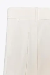 Pantalón blanco de tiro medio con pinzas delanteras y raya planchada. Confeccionado en hilatura con mezcla de algodón. Presenta bolsillos laterales, trabillas en la cintura y cierre frontal con cremallera, botón y gancho metálico.