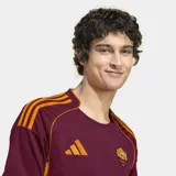 Camiseta de fútbol Adidas del AS Roma, modelo titular 25/26. Diseño color borgoña con detalles en naranja en cuello, mangas y hombros. Incluye logo de Adidas, escudo del club y tecnología Aeroready.