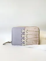 Billetera pequeña de cuero color lila y beige con cierre metálico y detalle de cordones cruzados en el frente.