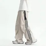 Pantalón deportivo color beige claro, estilo baggy o ancho, con cordón ajustable en la cintura y cierre completo impermeable (full zip) en los laterales. Presenta una franja vertical negra con tres líneas blancas a lo largo de la costura lateral, y un pequeño logo bordado en la parte superior del muslo.