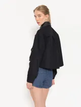 Chaqueta corta negra de gabardina combinada con cuero, con recortes delanteros, bolsillos plaqué con tapa y broches forrados. Presenta volados en denim.