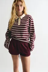 Remera tipo polo de manga larga con corte holgado, a rayas horizontales color borgoña y crema. Tiene cuello clásico color crema, tapeta con botones y hombros caídos.