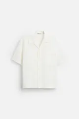 Camisa color blanco con textura de rayas verticales, corte relaxed fit, cuello bowling, manga corta, bolsillo de parche en el pecho y cierre frontal con botones.