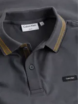 Chomba gris de piqué elástico con cuello polo y tapeta de dos botones. Presenta ribetes negros y dorados en cuello y mangas, parche con logo de Calvin Klein en el pecho y corte clásico.