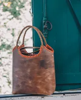 Bolso matero/cartera de cuero color marrón con detalles de costuras en hilo naranja. Tiene base rígida, manijas trenzadas y correa de cuero desmontable. Incluye accesorio sujetatermo y bolsillo para celular.
