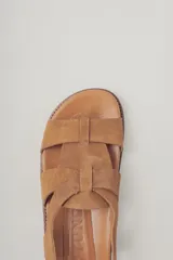 Sandalia plana de gamuza color marrón oscuro, con tiras cruzadas en la parte delantera y una tira fina ajustable con hebilla metálica en el tobillo. Posee plantilla de cuero y suela baja.