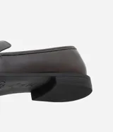Zapato mocasín de cuero color marrón oscuro con detalle metálico dorado en la parte delantera.