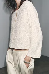 Chaqueta color crema de tejido de punto con lentejuelas, cuello barco, manga larga y cierre frontal con botones.