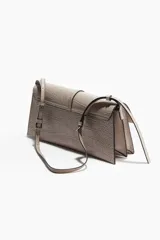 Cartera de hombro tipo sobre, color gris claro, con textura de piel de cocodrilo en relieve. Presenta una solapa frontal asegurada por una tira decorativa con hebilla metálica dorada. Incluye una correa ajustable fina para llevar al hombro.