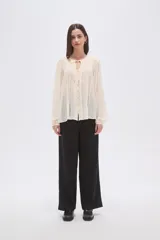 Blusa holgada color crema, confeccionada en viscosa, con frunces en el canesú superior y cuello en V con cordón ajustable terminado en mostacillas. Posee cierre frontal de botones y mangas largas abullonadas.
