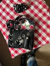 Cartera de cuero negra con correa regulable, estilo bandolera o al hombro. Decorada con charm de corazón metálico, encaje negro, cadenas y aros metálicos.