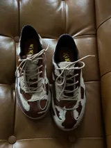 Sneakers de cuero con estampado de vaca en tonos marrón y blanco, con cordones beige y suela de goma color caramelo.