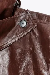 Chaqueta de Zara Woman Collection en tejido con acabado brillante efecto cuero. Presenta cuello solapa, manga larga con trabillas ajustables, bolsillos delanteros y cierre frontal con botones ocultos bajo solapa.