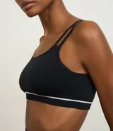 Top deportivo negro sin costuras, con escote asimétrico y doble tirante en los hombros. Presenta una delgada línea blanca horizontal en la banda inferior.