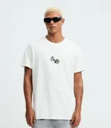 Remera blanca de algodón con cuello redondo y mangas cortas. Presenta un estampado estilo grafiti en la espalda con lettering en amarillo y rosa, y la ilustración de un perro con gorra y aerosol.