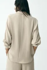 Camisa color marrón claro de cuello solapa y manga larga con trabilla y botón para remangar. Tiene un bolsillo de plastrón en el delantero, bajo asimétrico y cierre frontal con botones ocultos por solapa.