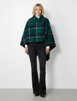 Capa corta de tejido grueso, color verde esmeralda con diseño de cuadros escoceses en azul marino y líneas finas en beige. Posee cuello alto que se cruza al frente y se cierra con un broche oscuro.