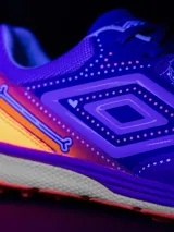 Championes de fútbol Umbro, color violeta con detalles en naranja y blanco. Presentan un diseño texturizado en la parte delantera y el logo de Umbro en los laterales. La suela es de color naranja con tapones para césped sintético.