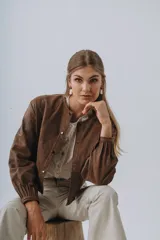 Campera bomber color beige de cuero, con cuello redondo, cierre frontal con botones a presión, mangas largas con puños elásticos y detalles de tachas metálicas en hombros y espalda.