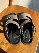 Sandalias bajas color beige con tiras cruzadas y hebilla al tobillo. Suela de goma negra con pespuntes blancos.