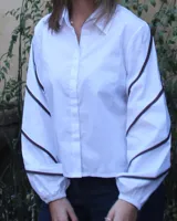 Camisa blanca de corte recto, con cuello clásico, cierre frontal con botones y mangas largas abullonadas con puños elásticos. Presenta detalles de ribetes marrones en mangas y hombros.