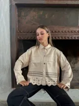 Camisa color beige con cuello clásico, cierre frontal con botones dorados, bolsillos de parche en el pecho y detalle de calado con terminación festoneada en el ruedo.