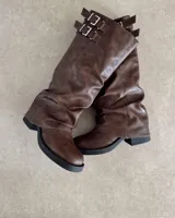 Bota alta de cuero vegano color marrón oscuro, con caña arrugada y detalle de dos hebillas ajustables en la parte superior. Posee suela y taco bajo de goma negra.