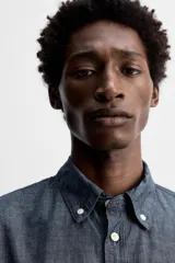 Camisa azul de algodón con textura chambray, corte regular fit, cuello abotonado, manga larga con puños abotonados, bolsillo de parche en el pecho y cierre frontal con botones.