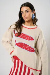 Sweater tejido color crudo con cuello redondo y mangas largas. Presenta dos peces rojos con detalles en azul bordados en el frente. Tiene ribete ondulado rojo en cuello, puños y ruedo.