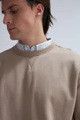 Sudadera de tejido liso color beige, con cuello redondo y puños y ruedo acanalados. Se lleva superpuesta a una camisa de cuadros celestes y blancos.