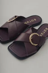 Sandalia plana de cuero, color marrón oscuro, con tira ancha cruzada sobre el empeine y hebilla metálica dorada.