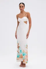 Vestido largo blanco con estampado tropical en tonos turquesa, naranja y marrón en el ruedo. Presenta escote corazón con detalle de nudo en el busto, recortes laterales en la cintura y tirantes finos con herrajes dorados.