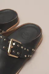 Sueco destalonado de cuero vacuno color negro, con hebilla ajustable dorada y detalles de tachas doradas.