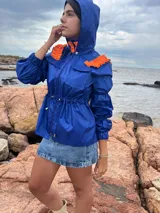 Piloto impermeable color azul eléctrico con detalles naranjas en los hombros, capucha integrada, cordón regulable en la cintura y mangas con volumen y puños elastizados.