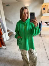Buzo tipo canguro oversize de tela plush, color verde brillante, con capucha y bolsillo canguro frontal. Presenta mangas largas con puños ajustados y cordones ajustables en los laterales inferiores.
