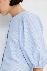 Blusa celeste y blanca a rayas verticales, de corte recto, con cuello redondo, cierre frontal con botones y mangas 3/4 abullonadas con puños elásticos.