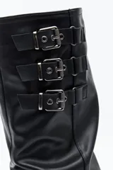 Bota de caña alta confeccionada en poliuretano con diseño de efecto fruncido y puntera redondeada. Presenta tres hebillas metálicas decorativas en el lateral superior de la caña y taco bajo.