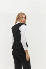 Chaleco blanco con finas rayas verticales negras, escote en V, cierre frontal con botones negros y volados en lino negro en los hombros. La parte inferior presenta otro volado en la misma tela del cuerpo.